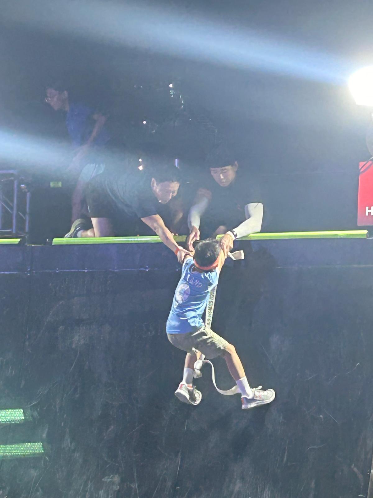 Two people reaching down to help a child over an obstacle wall