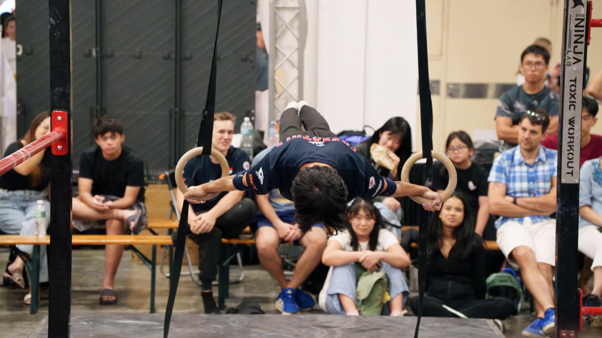 An athlete inverted on rings in front of a seated audience