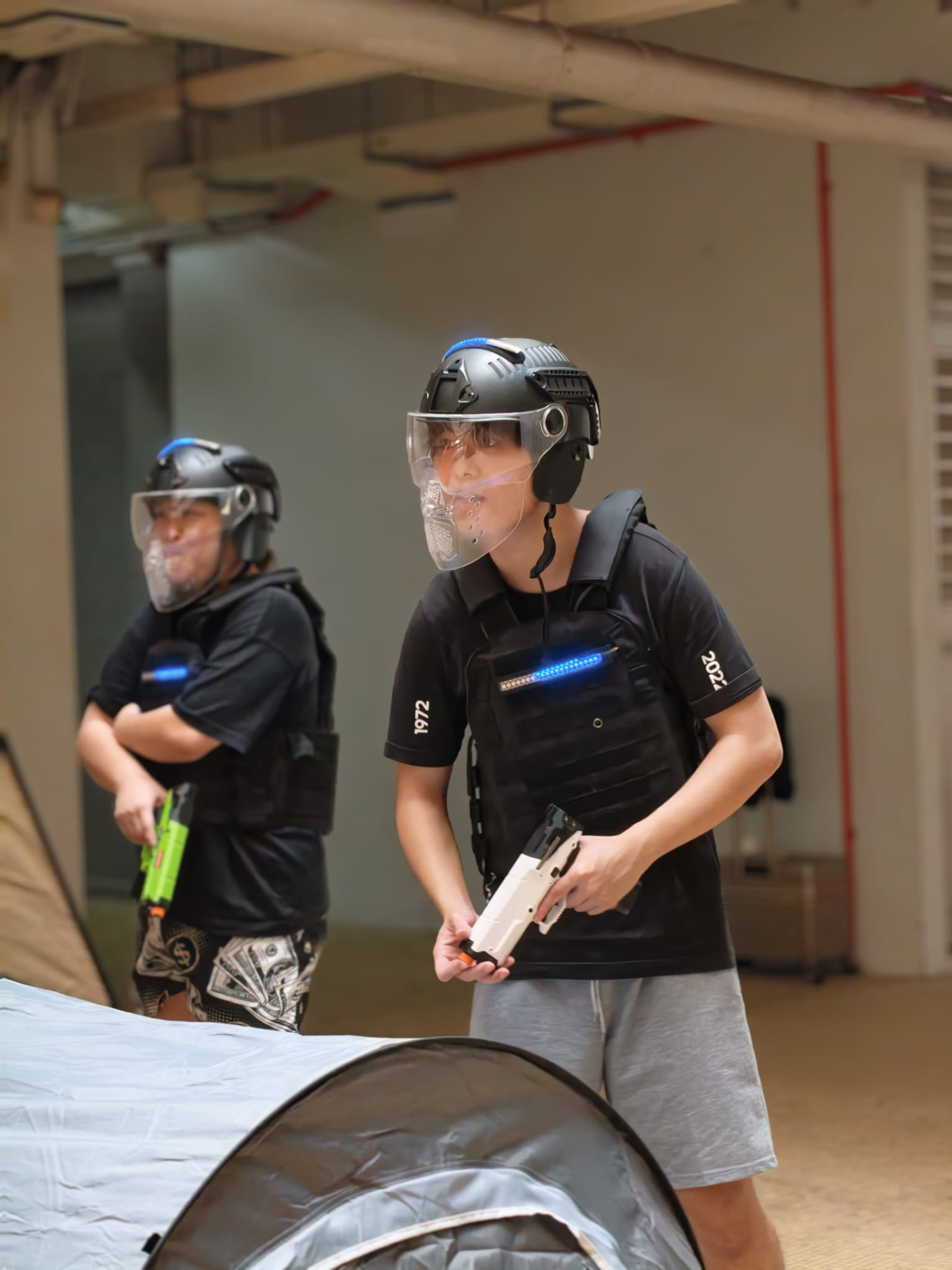 Two adult players wearing blue-lit hit-tracking vests and helmets, holding foam-dart blasters