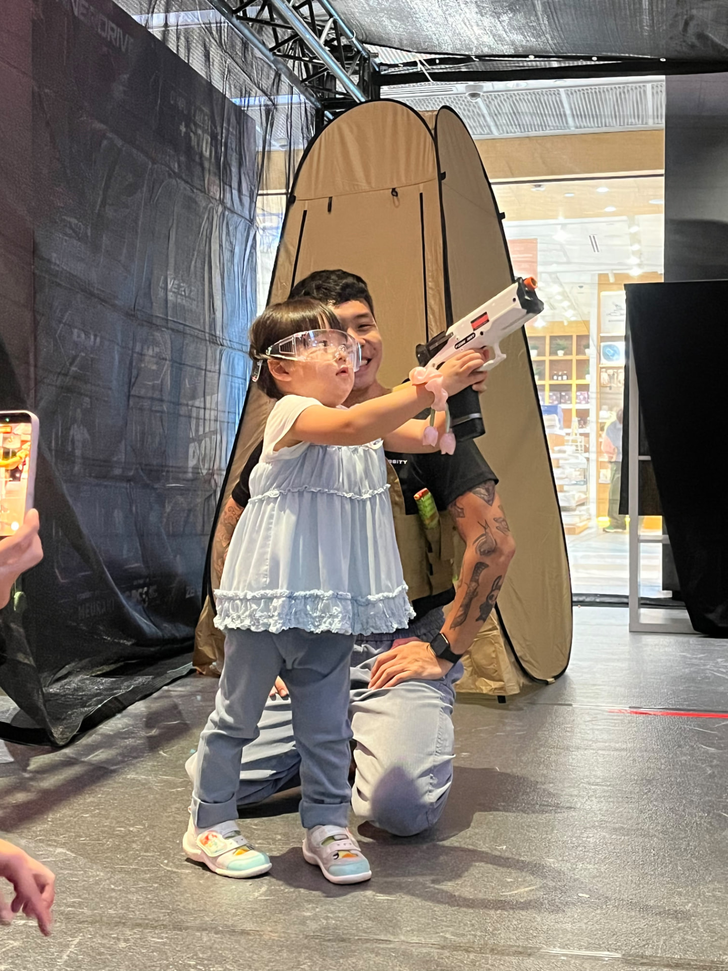A crew member kneeling behind a young girl in safety goggles as she takes aim.