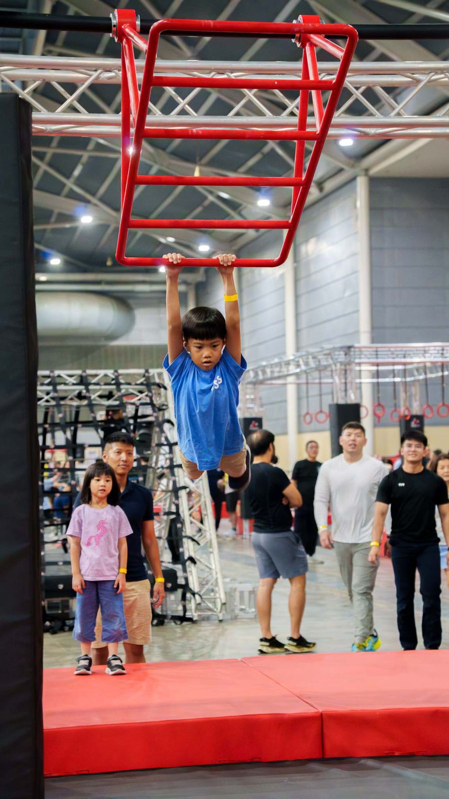A boy hanging from a red overhead ladder while families watch from the mats