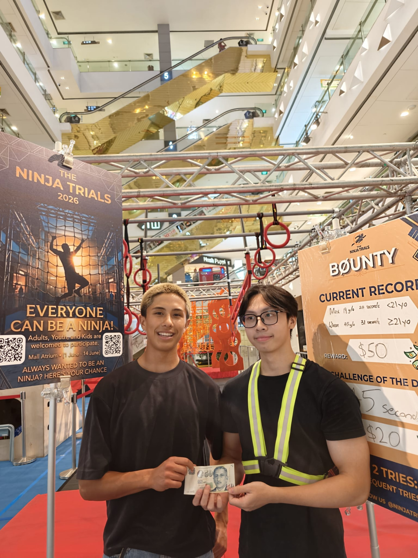 Two participants holding a fifty dollar note beside the Ninja Trials Bounty record board.